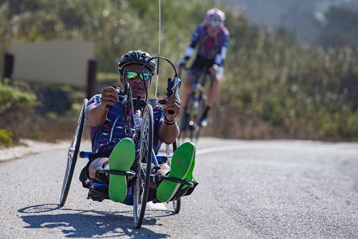 Two cyclists are on a road, focusing on the front cyclist in a recumbent handcycle with bright green shoes. Both are wearing helmets and sunglasses. The background shows trees and blurred scenery, suggesting speed and motion.