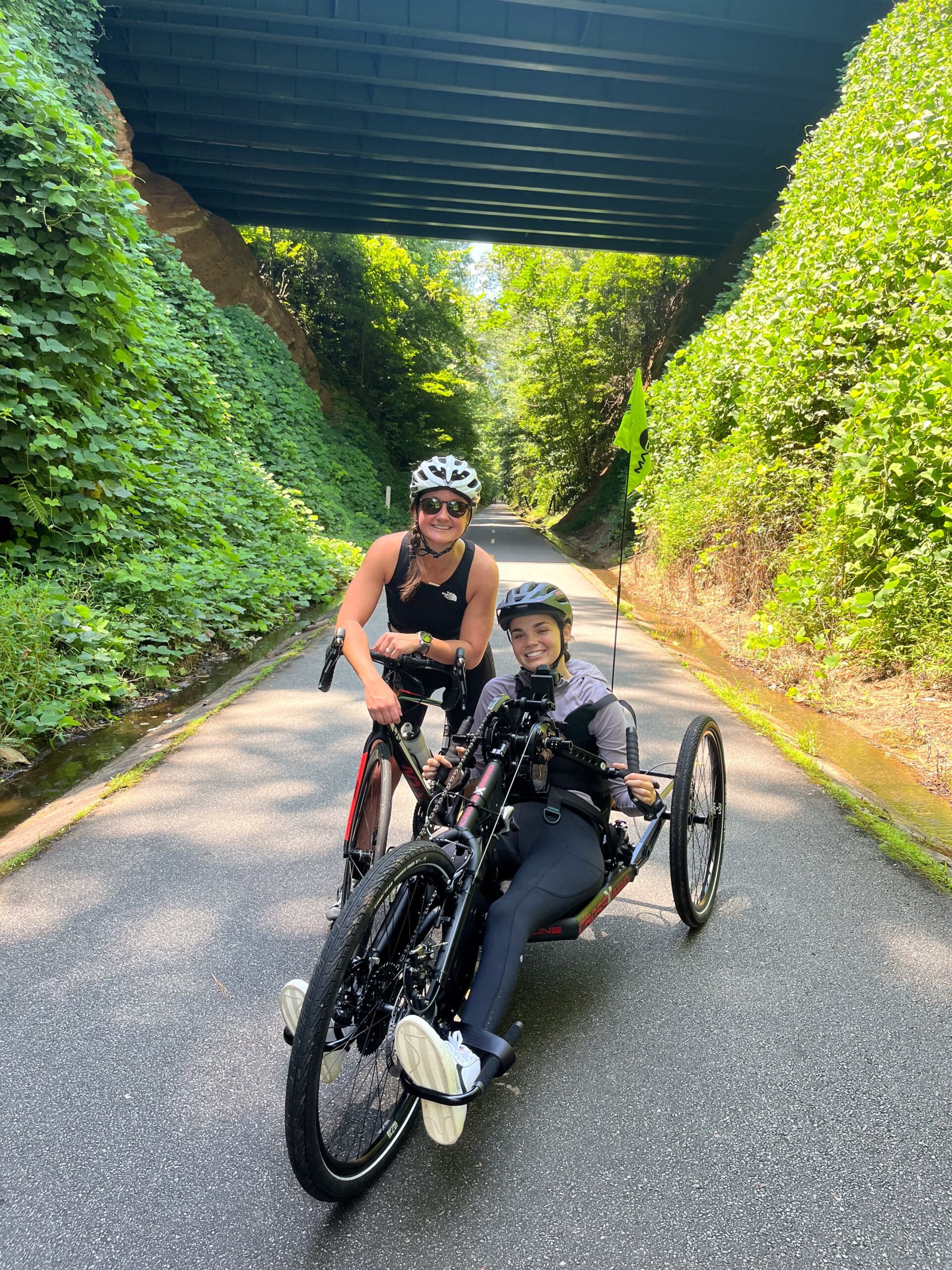 Two cyclists stop on a sunlit path under a bridge. One person is standing with a road bike, and the other is seated on a handcycle. Both are wearing helmets and smiling, surrounded by lush greenery.
