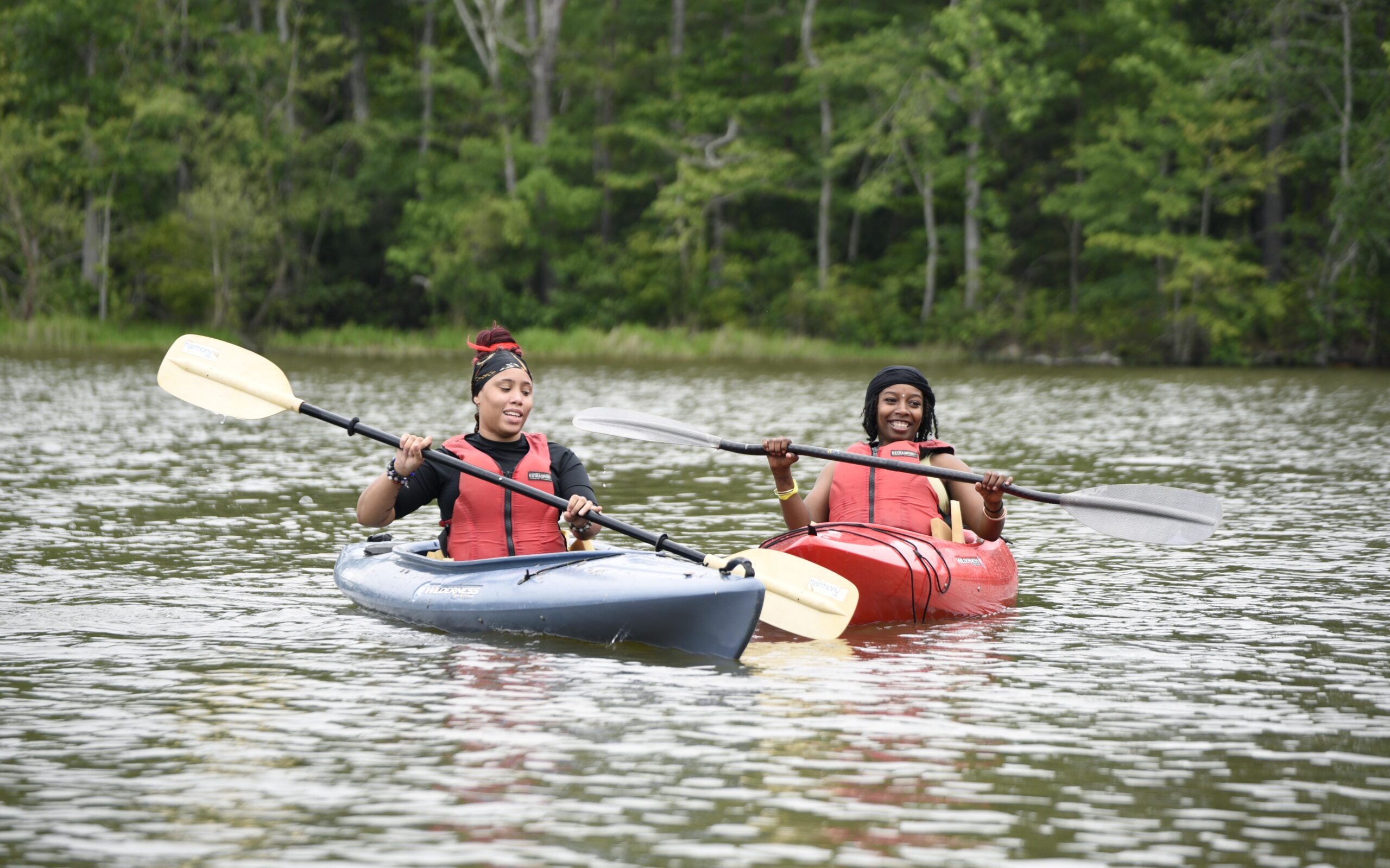 Two people kayaking on a calm lake surrounded by lush greenery. Both are wearing life vests and appear to be enjoying the activity. The kayaks are blue and red.