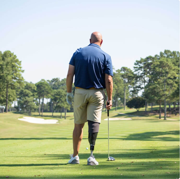 A man with a prosthetic leg, wearing a blue shirt and beige shorts, stands on a golf course holding a club. Lush green grass and trees are in the background under a clear blue sky.